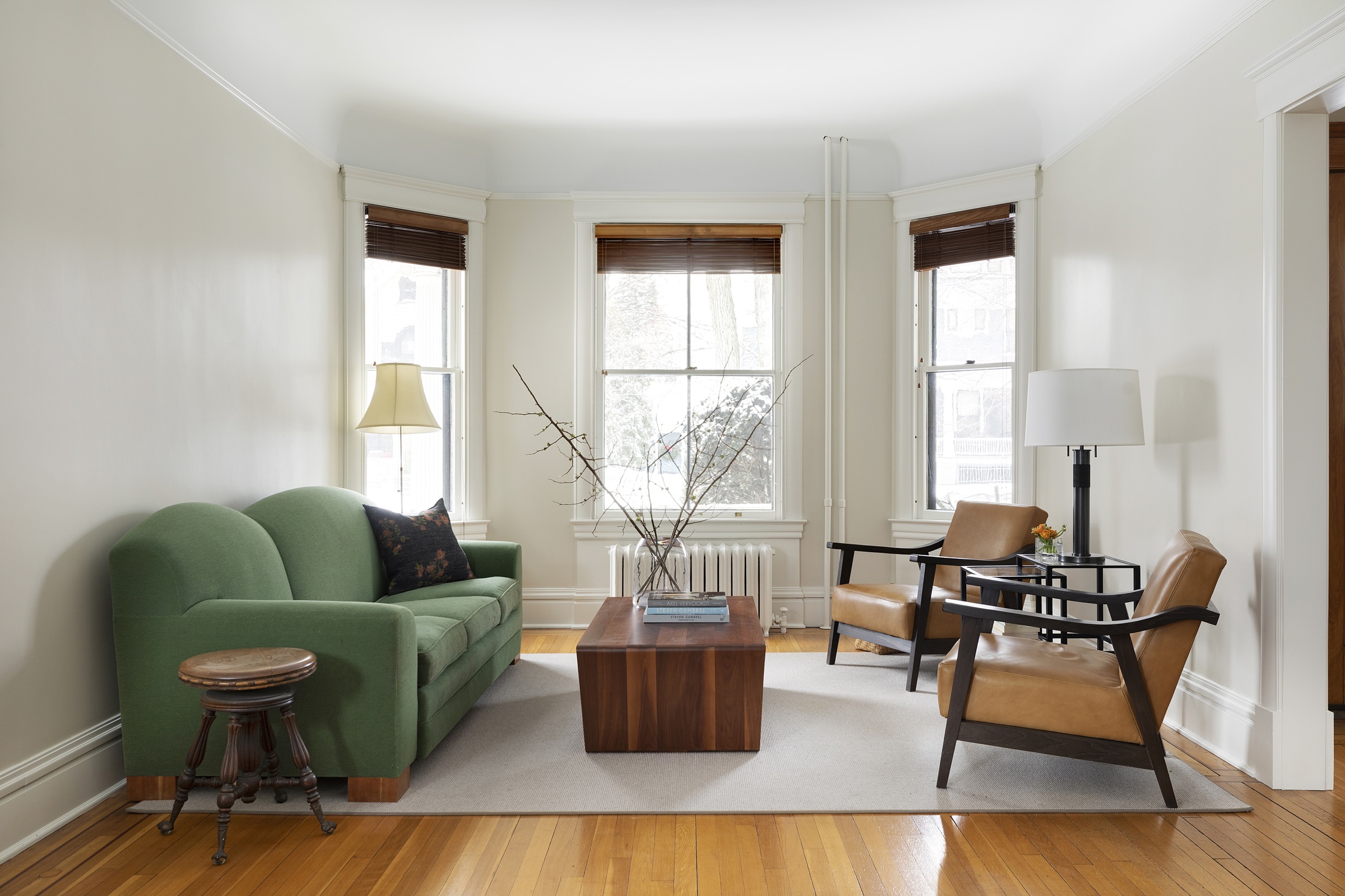 Bay-window sitting room with green sofa, walnut block coffee table and tan leather chairs on wood floors.