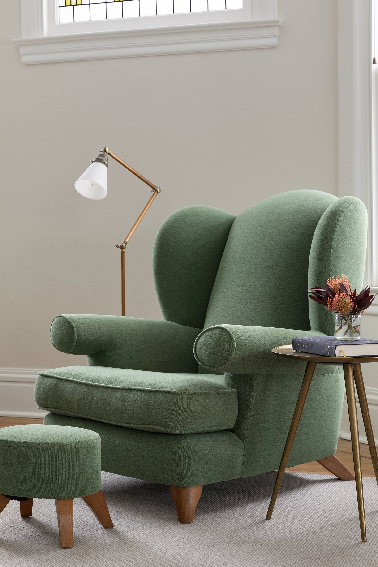 Green upholstered wing chair and ottoman beside a brass floor lamp and painted period woodwork.